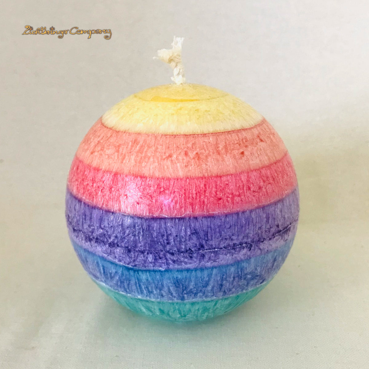 Regenbogenkerze in Kugelform, Farben von oben: gelb, orange, rot, lila, blau, grün. Jede Farbschicht ein anderer Duft. Vor hellem Hintergrund