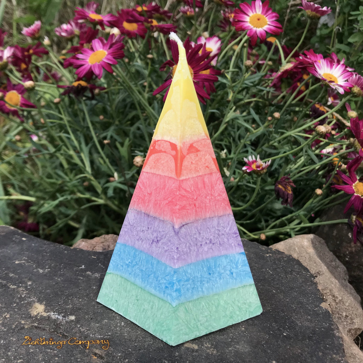 Regenbogenkerze in Pyramidenform, präsentiert auf einem Stein vor Blumen. Jede Farbschicht mit einem anderen Duft